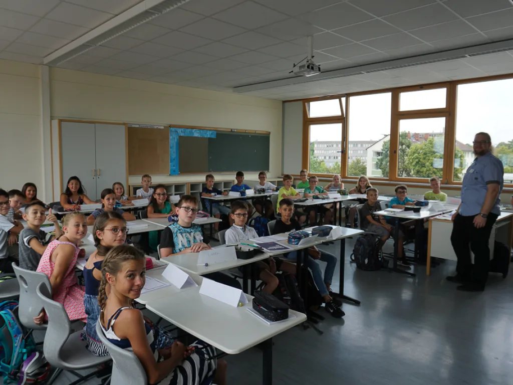 Kinder sitzen im Klassenraum, eine Lehrkraft steht daneben.